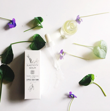 Load image into Gallery viewer, the elasticity serum bottle with some flowers and greens spread out on the ground around it