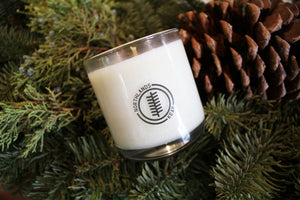 an up close video of a candle - the candle wax is a white color and is inside a clear glass container that doubles as a drinking tumbler once the candle burns out. the candle is sitting on pine sprigs and a pine cone