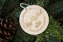 Load image into Gallery viewer, an up close look at a circular wooden ornament that says "Happiest of Holidays from Key West" it is laying on a pine wreath with a pine cone