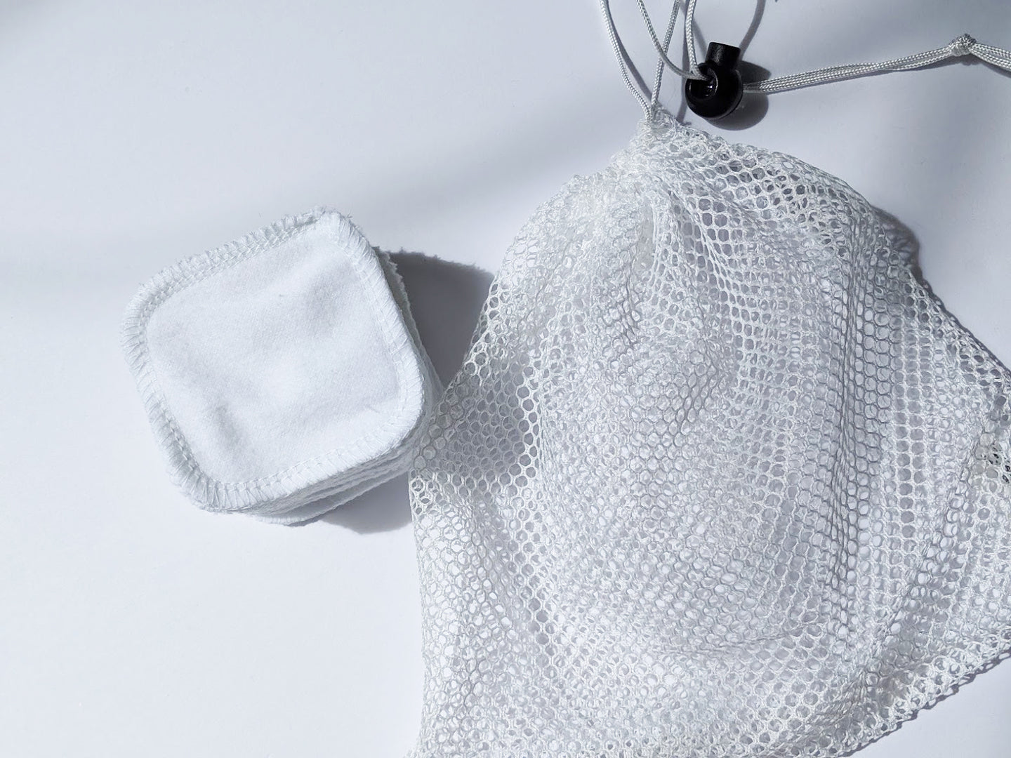 a mesh laundry bag next to a stack of facial rounds. The mesh laundry bag has a pull tab to be able to close fully.