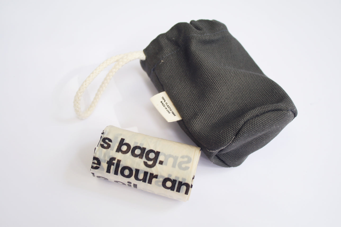 an up close look at a doggie poop bag holder next to the roll of baggies that go inside. The pop bag holder is a gray color. It has a soft cotton handle and a small tag on the side that says 