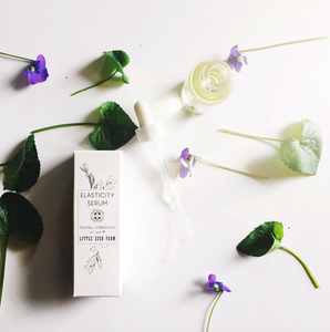 the elasticity serum bottle with some flowers and greens spread out on the ground around it