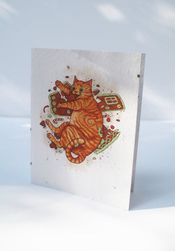 a plantable seed card - the card has a textured look from the seeds imbedded in the paper. There is a drawing of an orange cat eating a gingerbread man and gingerbread house on the front of the card. The inside of the card is blank.