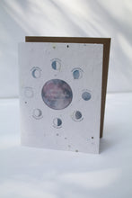 Load image into Gallery viewer, a plantable seed card - the card has a textured look from the seeds imbedded in the paper. There is a drawing of the moon phases on the card in an indigo color. The moon phases are surrounding one large moon in the center that reads &quot;wishing you many more moons&quot;
