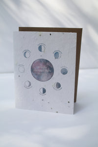 a plantable seed card - the card has a textured look from the seeds imbedded in the paper. There is a drawing of the moon phases on the card in an indigo color. The moon phases are surrounding one large moon in the center that reads "wishing you many more moons"
