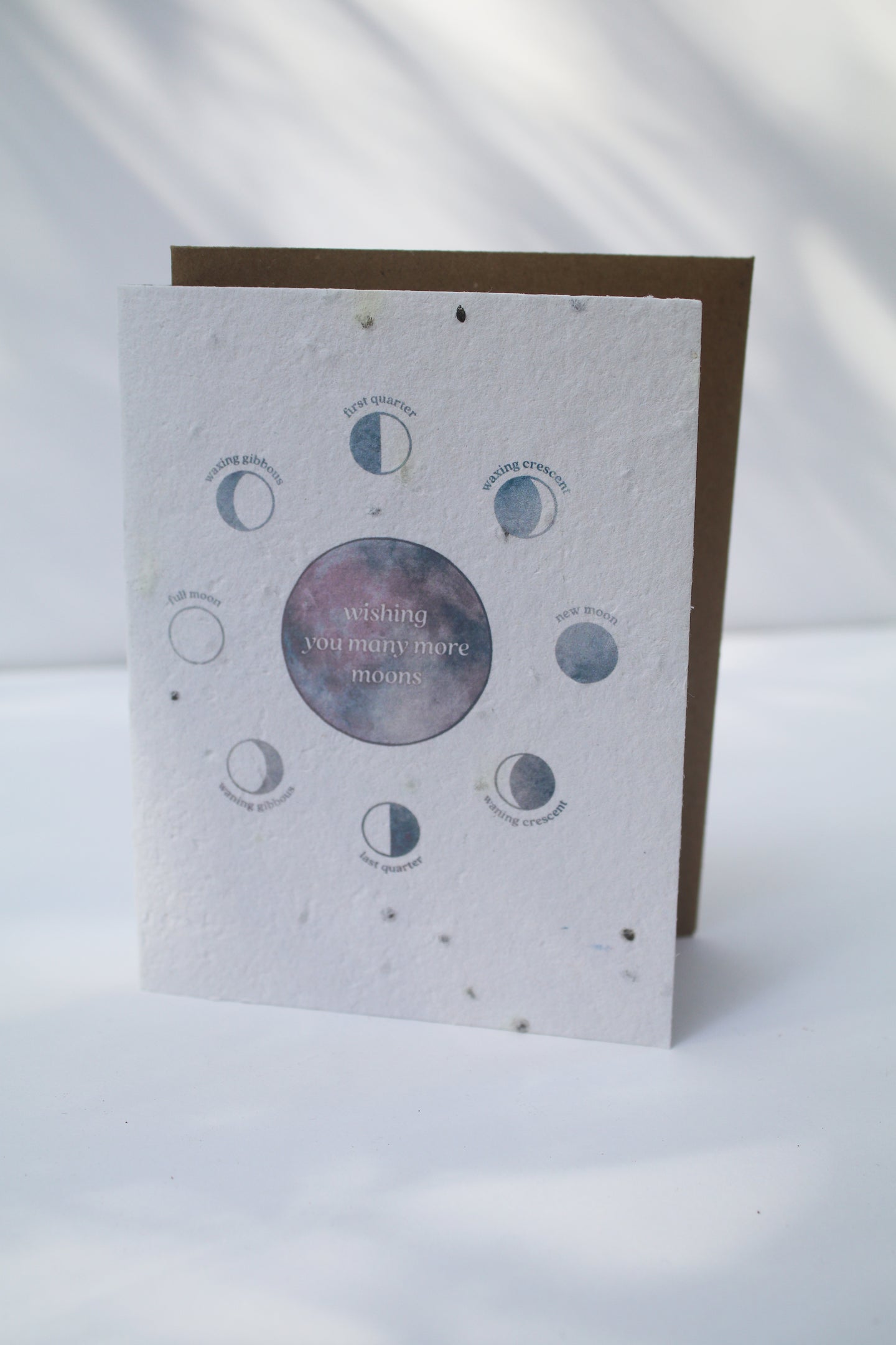 a plantable seed card - the card has a textured look from the seeds imbedded in the paper. There is a drawing of the moon phases on the card in an indigo color. The moon phases are surrounding one large moon in the center that reads 