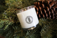 Load image into Gallery viewer, an up close video of a candle - the candle wax is a white color and is inside a clear glass container that doubles as a drinking tumbler once the candle burns out. the candle is sitting on pine sprigs and a pine cone
