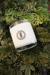 an up close video of a candle - the candle wax is a white color and is inside a clear glass container that doubles as a drinking tumbler once the candle burns out. the candle is laying on pine sprigs.