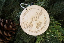 Load image into Gallery viewer, an up close look at a circular wooden ornament that says &quot;Happiest of Holidays from Key West&quot; it is laying on a pine wreath with a pine cone
